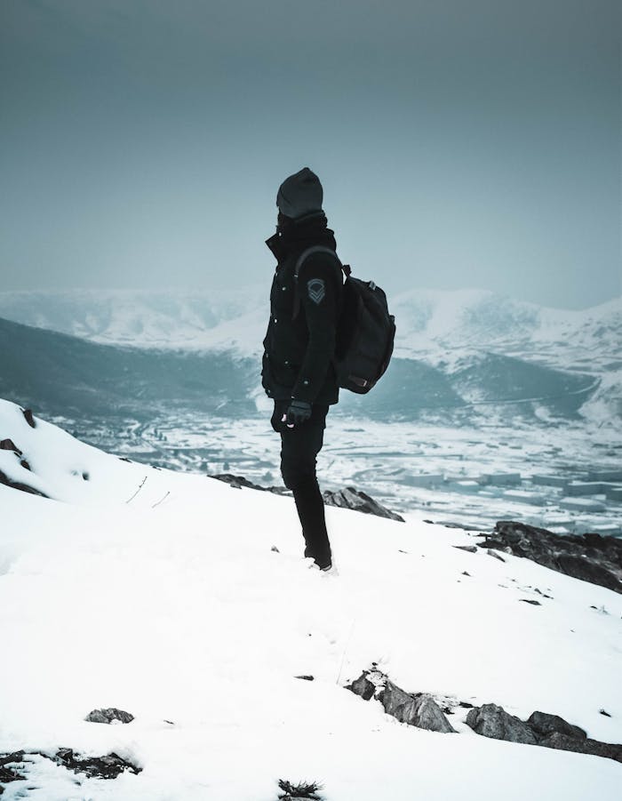A lone hiker stands in winter snow, exploring a vast mountain vista.