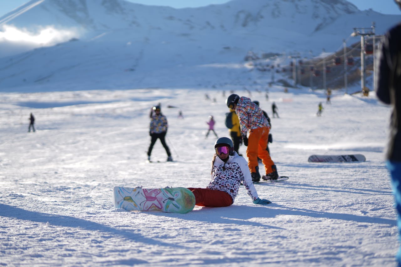 A snowboarder sitting on a snowy slope, enjoying the sunny winter day on a busy ski resort.