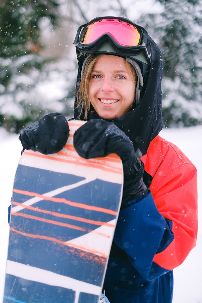 A cheerful woman in winter gear holds a snowboard, enjoying the snowy outdoors.